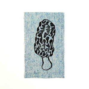 Puede incluir: Ilustración en blanco y negro de una seta de colmenilla impresa sobre un pequeño trozo rectangular de tela vaquera azul claro. La seta tiene un sombrero texturizado en forma de esponja y un tallo delgado. La tela tiene un borde deshilachado.