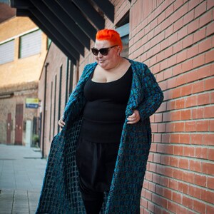 Op de afbeelding: Een vrouw met fel oranje haar draagt een blauw-zwart gepatroneerd vest over een zwarte tanktop en een zwarte broek. Ze staat voor een bakstenen muur.