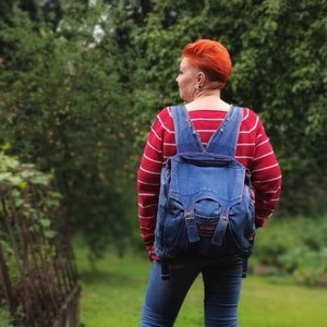 May include: A woman with bright red hair is wearing a red and white striped shirt and a blue denim overall dress. She is carrying a blue denim backpack and wearing black cowboy boots. She is standing on a path in a grassy area with trees in the background.