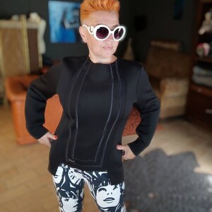 May include: A woman with short, bright orange hair is wearing a black long-sleeved jacket with white stitching and black and white leggings with a graphic print of faces. She is wearing white sunglasses.