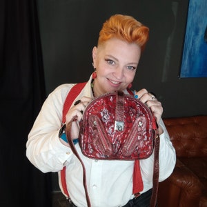 May include: A woman is holding a red leather crossbody bag with a textured pattern and a silver lock. The bag has two zipped pockets on the front.