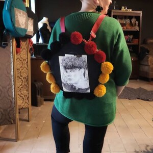 May include: A green sweater with a black and white photo of a person on a round, black bag with yellow and red pom-poms. The person is wearing a black shirt and has short, dark hair.