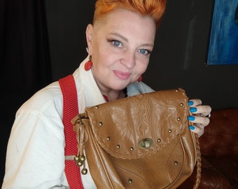 May include: A woman with short, bright red hair is holding a brown leather satchel with gold accents and a braided strap. She is wearing a white button-down shirt and red suspenders.
