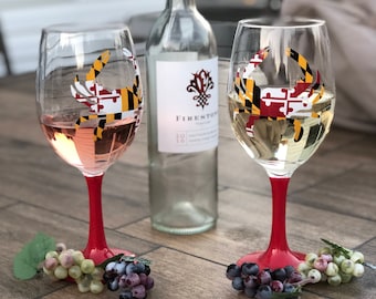 May include: Two wine glasses with red stems and Maryland flag designs, filled with rosé and white wine, sit on a wooden table. A clear wine bottle and grapes are also present. The glasses are decorated with the Maryland flag.
