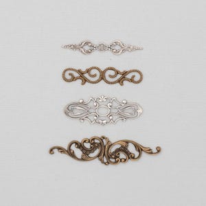 May include: Four ornate metal embellishments in silver, bronze, and gold tones are arranged on a light gray background. The decorative pieces feature intricate scrollwork and symmetrical designs, suitable for craft projects.