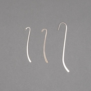 May include: Three minimalist, curved metal earrings in silver and gold tones are displayed against a gray background. The earrings have a hook design, with a slender, elongated shape. The earrings are likely for pierced ears.