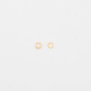 May include: Two gold-colored metal jump rings of different sizes, each featuring a small opening in the circular band. These jewelry findings are circular in shape and have a smooth, polished finish, presented side by side on a plain surface.