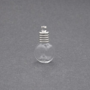 May include: A small, clear glass vial pendant with a round, bulbous body and a silver-toned metal cap featuring horizontal ridges and a small circular loop at the top for attachment to a necklace or other jewelry component.
