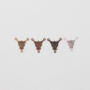May include: Four decorative metal jewelry connectors in bronze, copper, dark bronze, and silver. Each connector has a triangular shape with three loops and a central decorative design. The connectors are arranged horizontally against a white background.