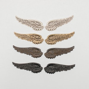 May include: A collection of decorative metal wing pairs in silver, gold, bronze, and black. Each wing pair features detailed feather textures, perfect for crafting or embellishing projects. The wings are arranged on a white background.