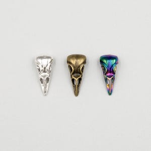 May include: Three bird skull pendants in silver, bronze, and iridescent colors. The pendants are detailed with eye sockets and a pointed beak, perfect for jewelry making or crafting projects.