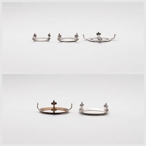 May include: A collection of silver and gold-toned metal crown-shaped objects, each with a circular base and ornate fleur-de-lis or cross detailing. The crowns vary in size and design, set against a white background.