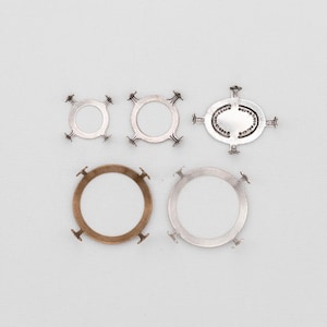 May include: An assortment of metal jewelry findings in various shapes and sizes. The collection includes circular and oval frames with decorative accents and mounting posts. The metal finishes range from silver to bronze, offering a variety of design options.