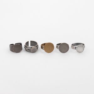 May include: Five adjustable rings with circular settings in various metallic finishes. The rings include dark gray, silver, and bronze tones. The settings are flat and round, suitable for adding custom designs or embellishments. The rings are displayed against a white background.