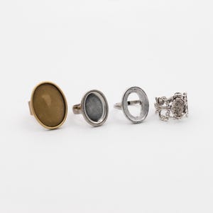 May include: Four rings with different designs. One ring has a bronze-colored oval setting, another has a silver-colored oval setting, and a third has a silver-colored open oval setting. The fourth ring has a detailed, ornate silver design with a central stone.