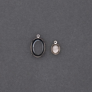 May include: Two silver-toned oval pendant settings, one large and one small, with a loop at the top for attaching to a chain or other jewelry component. The settings have a dark interior and a raised edge.