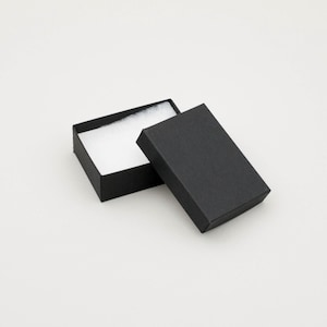May include: A small, open black gift box with a white, fluffy interior. The lid is slightly ajar, revealing the box's contents. The box is rectangular and sits on a white surface, suitable for jewelry or small gifts.