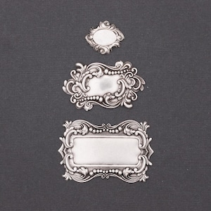 May include: Three silver-coloured metal embellishments with ornate designs. The largest is rectangular, the middle one is oval, and the smallest is a small oval shape. Each has detailed scrollwork and a blank centre for personalisation.