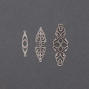May include: Three silver filigree jewellery components. The pieces vary in size and design, including an elongated diamond shape, a teardrop shape, and a rectangular shape with ornate scrollwork. They are displayed against a dark grey background.