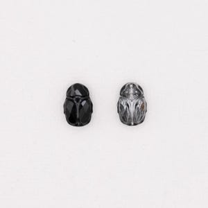May include: Two scarab beetle figurines. One is black and faceted, the other is clear with detailed carvings. Both are set against a white background. The beetles are likely decorative items or jewelry components.