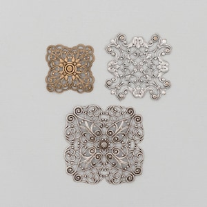 May include: Three ornate, square metal filigree embellishments. One is gold-toned with a central circular design, and the other two are silver-toned with intricate floral and scroll patterns. The embellishments are displayed on a light gray background.
