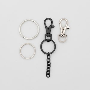 May include: An assortment of keychains and key rings. The keychains include a black swivel clasp with a key ring and chain, and a silver swivel clasp. Two silver key rings are also included.