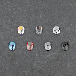 May include: Seven faceted skull-shaped embellishments in various colours, including iridescent, silver, blue, red, grey, and black. The skulls have detailed facial features and are arranged on a dark grey surface.