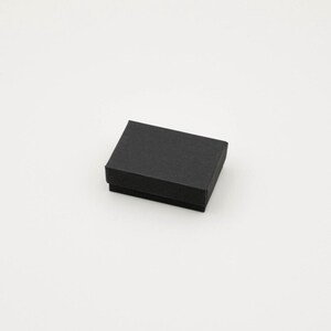 May include: A small, rectangular black box with a lid, likely for jewelry or small gifts. The box is made of a matte material and sits on a white surface. The box is empty.