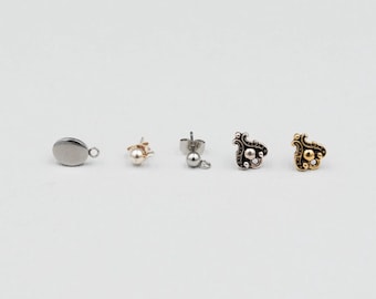 May include: An assortment of earrings in various styles and finishes. The collection includes a silver oval charm, a gold-toned stud with a small ball, a silver stud with a dangling ball, and two ornate stud earrings in silver and gold tones.