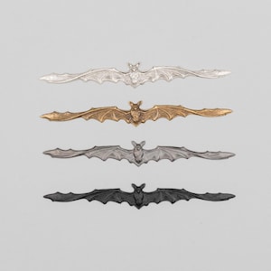 May include: Four metal bat-shaped decorative accents in silver, gold, gray, and black. Each bat has outstretched wings and a detailed body. The bats are arranged horizontally against a light gray background, suitable for Halloween or gothic decor.