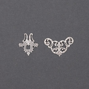 May include: Two silver-colored filigree embellishments. One is a symmetrical design with a central teardrop shape and dotted edges. The other is a curved, ornate piece with swirling patterns and a central floral element. Both are set against a dark gray background.