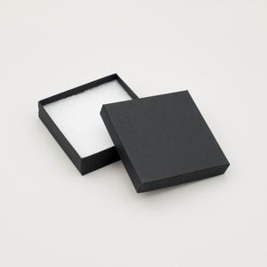 May include: An open, black square gift box with a white, padded interior. The lid is slightly ajar, revealing the inside. The box is on a white background. Ideal for jewelry or small gifts.