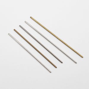 May include: Five metal knitting needles in varying colors: silver, bronze, and gold. The needles are arranged diagonally against a white background, showcasing their smooth, rounded tips and slender shafts. The needles are used for knitting.