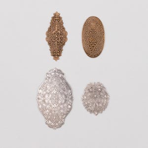 May include: Four ornate metal embellishments in gold and silver tones. The designs include floral and scroll patterns, with oval and elongated shapes. The pieces are likely for crafting or decorative purposes.