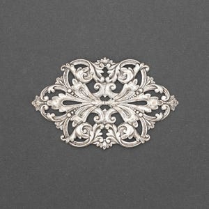 May include: A silver-colored, oval-shaped decorative element with an ornate, symmetrical design. The piece features intricate floral and scrollwork patterns, creating a vintage or antique aesthetic. The background is a dark gray.