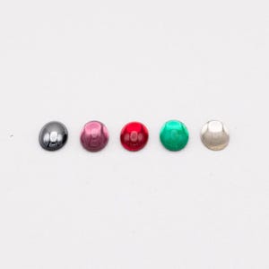 May include: Five colorful, domed cabochons in a row. The colors are, from left to right, dark gray, pink, red, green, and silver. The cabochons are smooth and shiny, with a rounded shape.