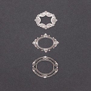 May include: Three ornate, silver-colored metal frames with open centers are arranged vertically against a dark gray background. The frames feature intricate scrollwork and decorative details, showcasing a vintage or antique aesthetic. The shapes include a hexagon, an oval, and another oval.