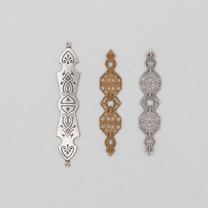 May include: Three decorative metal pendants in silver and gold. The silver pendant on the left has an ornate design with a loop at the top. The gold pendant in the centre has a geometric pattern. The silver pendant on the right has a floral design.