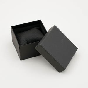 May include: An open black watch box with a textured exterior and a black interior. The lid is slightly ajar, revealing a watch resting on a black cushion. The box is square and sits on a white surface.
