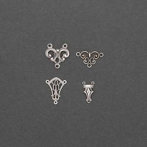 May include: Four silver-coloured metal jewellery findings in various ornate designs. The findings include filigree and scrollwork patterns, with loops for attaching to other components. The background is a dark grey.