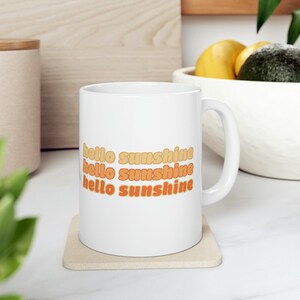 May include: White ceramic mug with the text "hello sunshine" repeated three times in a gradient of orange and yellow.