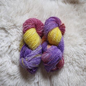 Puede incluir: Dos ovillos de hilo en una combinación de colores morado, amarillo y rosa. El hilo es suave y esponjoso, y es perfecto para tejer o hacer ganchillo.