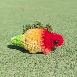 Puede incluir: Un juguete de dinosaurio hecho a mano con ganchillo en un degradado de colores: verde, amarillo, naranja y rojo. El dinosaurio tiene un pequeño ojo negro y una espina dorsal verde texturizada. El juguete está sobre una superficie verde.