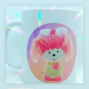 May include: White ceramic mug with a pink and white cartoon dog wearing glasses and a pink bow. The dog is on a pink, orange, and purple background. The mug has a handle and the text "@prettyglitchness" is at the bottom.