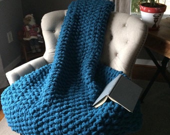 May include: A teal blue chunky knit throw blanket draped over a white armchair with a book on top.