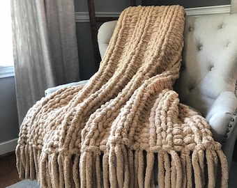 May include: A chunky knit throw blanket in a solid beige color, featuring a thick, textured yarn construction with vertical ribbed patterns and long, thick fringe tassels hanging from the bottom edge, draped over a tufted upholstered chair.