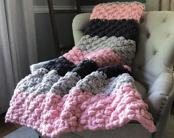 May include: A pink, gray, and black chunky knit throw blanket draped over a gray armchair. The blanket is made with large, fluffy yarn and has a striped pattern.