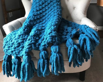 May include: A teal blue knitted throw blanket with tassels on a white armchair. The blanket is made with a chunky yarn and has a textured, bobbly appearance.