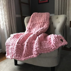Può includere: Una coperta di maglia rosa spessa drappeggiata su una poltrona grigia chiaro. La coperta è realizzata con un filato spesso e ha una texture morbida e soffice.