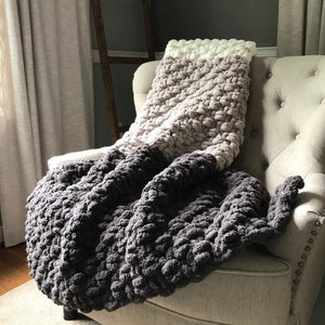 May include: A gray, pink, and white chunky knit throw blanket draped over a beige armchair. The blanket is made of thick yarn and has a textured, puffy appearance.